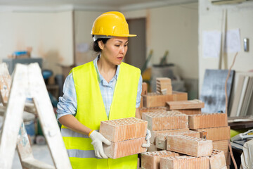 Asian female builder in a yellow vest and hardhat carrying hollow clay bricks and putting them on brick stack