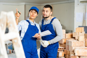 Two focused woman and man builders in blue coveralls discussing workflow while standing at...
