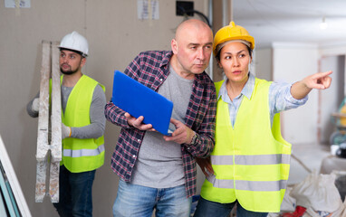 Caucasian man architect with laptop and asian woaman engineer discussing work while standing inside apartment during repair works. Woman pointing finger.