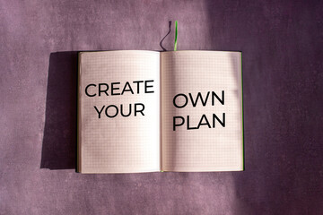 An open notebook is on the gray background. The word "Create your own Plan" is written on a blank sheet of notebook. Learning, education concept.