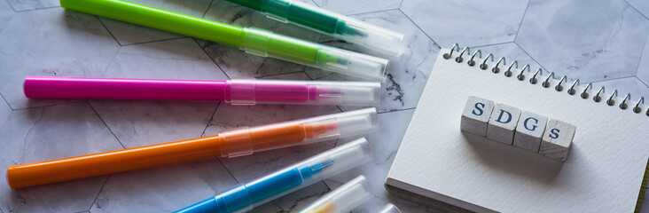 There is small sketchbook and color pens on the stone table with word cube formed SDGs.