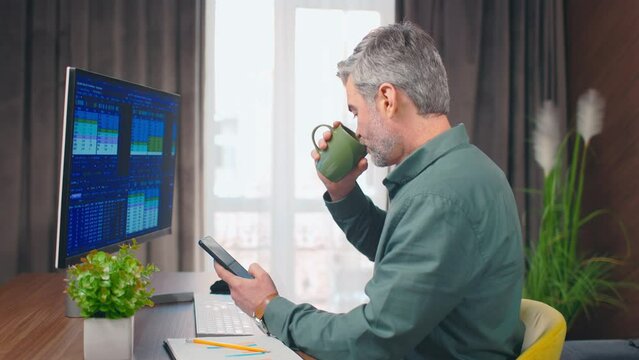 Side view close up portrait on middle aged caucasian man scrolling phone, checking social media and drinking coffee. Stock graphs image on computer. Office concept.