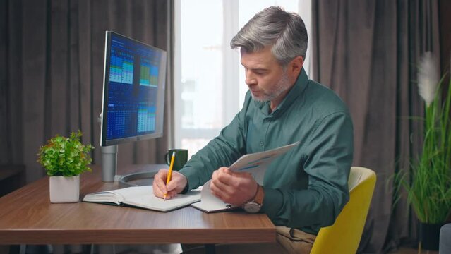 Side view close up portrait on handsome middle aged caucasian man writing in journal, checking stock market graphs. Stock graphs image on computer. Office concept.