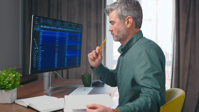 Side view close up portrait on handsome middle aged caucasian man studying cryptocurrency, writing in journal, checking stock market graphs. Stock graphs image on computer. Office concept.