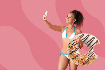 Beautiful young African-American woman with big donut taking selfie on pink background