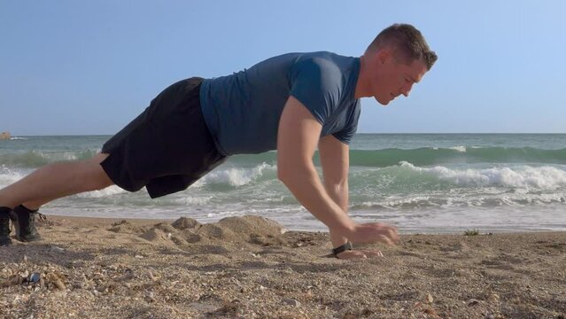A young Caucasian man with short hair doing push-ups (sports) on the sand beach by the sea with his connected watch