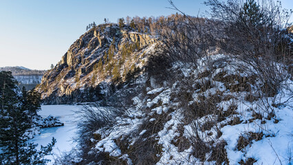 A picturesque cliff against the blue sky. Trees grow on the rocky slopes. Dry grass on snow-covered ground. At the foot are the bends of a frozen river. Altai.  Katun. Patmos