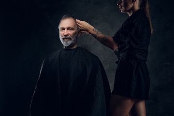 Portrait of woman hairdresser cutting hairs of her elderly customer against dark background.