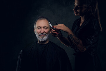 Portrait of female barber cutting hairs of her joyful elderly customer against dark background.