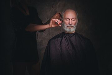 Studio shot of bearded senior man and female barber with scissors against dark background.