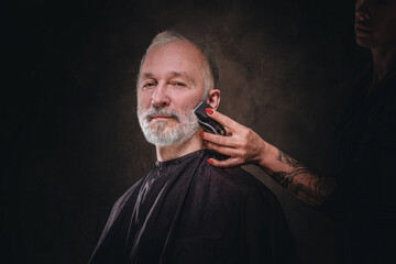 Portrait of female hairdresser with tattooes cutting stylish old man against dark background.