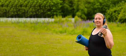 Plus size white Caucasian woman in headphones, a black T-shirt with a yoga mat in her hands is...