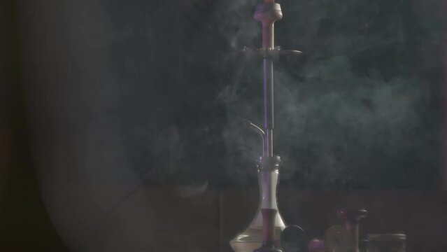 Smoke is swirling around the hookah on the dark background, slow motion