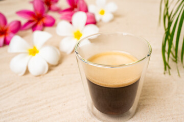 Double Wall Glass Espresso On Sand Beach Next To Frangipani Flowers.