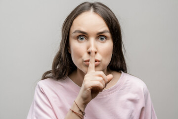 The girl shows her finger at her lips. A sign of silence. Brunette girl with long hair on a gray background