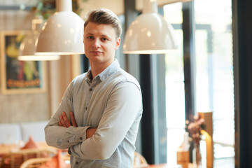 Portrait of content handsome young male restaurant manager in shirt crossing arms on chest in contemporary food establishment