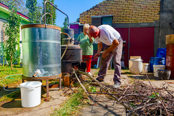 Woman is manually mixing fruit marc in distillation apparatus, man is throwing firewood for...
