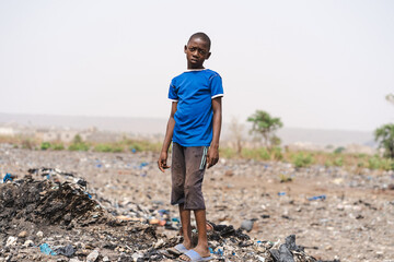 African child with slumped shoulders stands sadly in the middle of a garbage field, symbolizing the...
