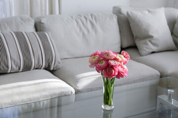 Close up shot of glass coffee table with bouquet of beautiful bicolor ranunculus flowers in a vase on foreground and gray textile couch on the background. Copy space, close up, natural light.