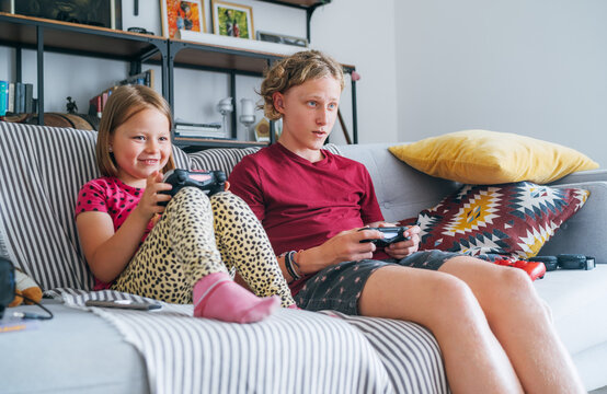 Smiling little Sister with brother sitting on sofa in living room, looking TV and playing console video game using wireless controllers. Careless childhood, modern entertainment technologies concept.