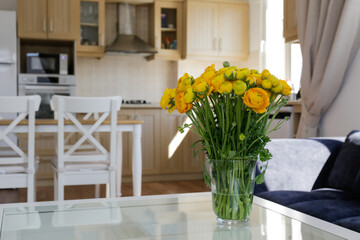 Bouquet of yellow ranunculus flowers in glass vase on the kitchen table. Stylish modern kitchen with small beige tile and wooden cupboard. Close up, copy space, background.