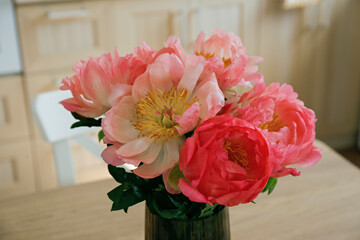 Beautiful peony flowers in glass vase on the kitchen table. Close up shot of a lush bouquet in stylish modern kitchen with small beige tile and wooden cupboard. Close up, copy space, background.