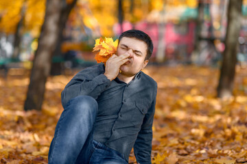 portrait of a man sitting in a glade with yellow maple leaves and scratching his nose, allergy...