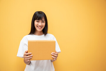 Fototapeta na wymiar Happy Asian woman smiling and holding package parcel box isolated on yellow background, Delivery courier and shipping service concept. Service mind