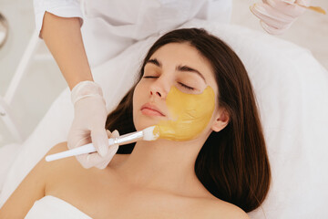 The cosmetologist applies a facial gold mask to the woman's face. Cosmetology and facial skin care in beauty salon. Gold mask. Cosmetic procedure. 