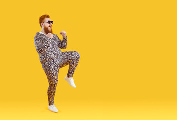 Funny fat redhead man in animal print pajama dancing on yellow studio background. Smiling overweight red-haired guy in cool suit perform winner dance celebrate success. Copy space.
