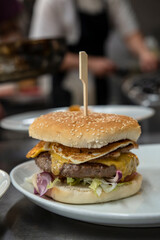 Restaurant kitchen imagery with a freshly prepared cheeseburger with toasted sesame bun, fresh vegetables, beef burger, melted cheese and fried egg resting on a plate and a busy chef in the background