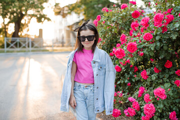 Smiling cute kid girl 6-7 year old wear mom denim jacket and sun glasses posing over pink rose flowers over nature background. Childhood. Summer season.