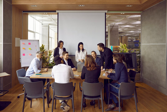 Cheerful And Smart Colleagues Are Discussing Their New Business Project At Brainstorm In Office. Group Of Business Men And Women Sitting At Conference Table In Modern Office Center With Loft Interior.