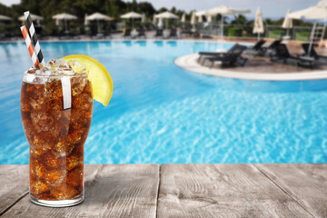Tasty refreshing soda drink with ice cubes on wooden table near outdoor swimming pool, space for...
