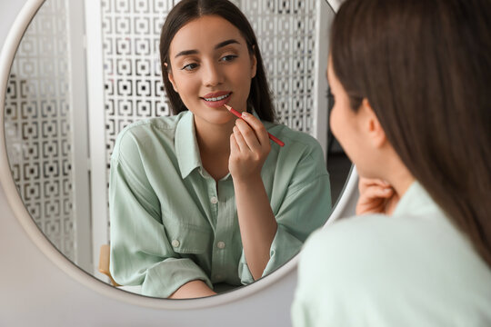 Beautiful young woman applying cosmetic pencil on lips near mirror indoors