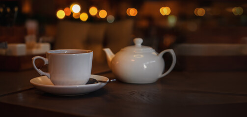 White cup with teapot. Pot standing on saucer in soft focus on naturally blurred background. Coffee, tea house, bokeh lights. The concept of a cozy pastime, tea ceremony