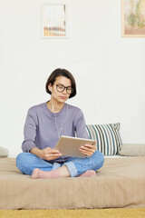 Fototapeta premium Serious student girl with short brown hair sitting with crossed legs on bed and using tablet in earphones