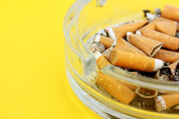 Lots of dirty used cigarette butts in an ashtray on a yellow background. Glass ashtray. Health, smoking, harm to health, Cancer of the lungs. View from above. Copy space for text