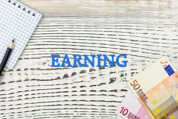 Naklejka premium EARNING - word (text) and euro money on a white wooden table, notebook, notepad. Business concept: buying, selling, commerce (copy space).