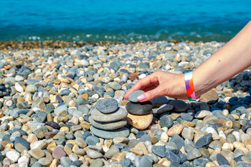 Female hands build balanced pebbles pyramid on beach against blue sea on sunny day. Stone cairn outdoors. Zen balance stones, meditation, spa and harmony concept