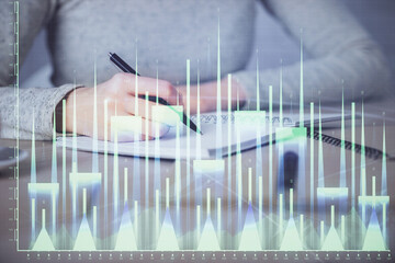 Hands writing information about stock market in notepad. Forex chart holograms in front. Concept of research. Multi exposure