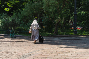A lonely elderly Polish nun in a gray robe, carrying a suitcase on wheels and a bag over her...