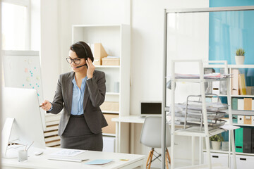 Content confident female manager in gray jacket standing at desk and discussing project while video conferencing with colleagues