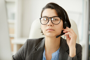 Portrait of serious confident beautiful female technical support operator in eyeglasses wearing headset with microphone for communication with clients