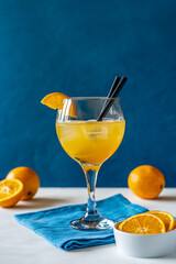 Yellow bird cocktail with rum, orange and lime juice, oranges, blue napkin. White table. Dark blue background. 