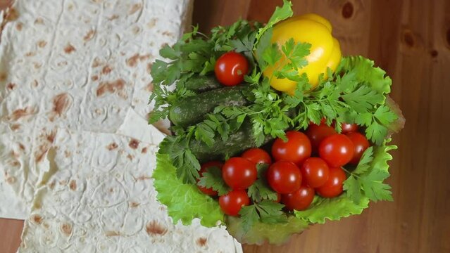 Raw vegetables red cherry tomatoes, small cucumbers, yellow bell pepper, fresh parsley and lettuce leaves on a rotating plate against the background of a tortilla lying on a wooden table top 