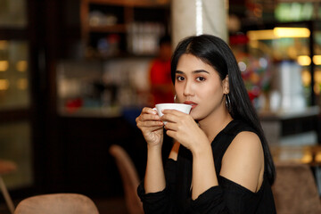 A young woman with long hair, black dress, tan skin, sitting at a table with a cup of coffee in a coffee shop.