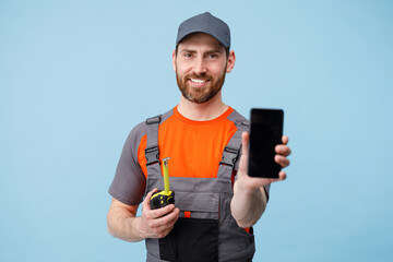 Smiling Man builder in uniform holding tape measure and showing empty screen phone in hands 