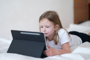 Little girl carefully looks at tablet while lying on bed