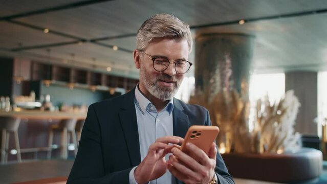 Portrait of good-looking thoughtful middle-aged Caucasian man looking away, writing message. Smiling Caucasian restaurateur chatting, using smartphone while sitting on couch in comfortable restaurant